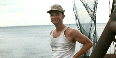 Movie gif. Tom Hanks as Forrest Gump stands on a boat with a wide smile on his face. His eyes beam as he gazes ahead, raising a hand and waving energetically.